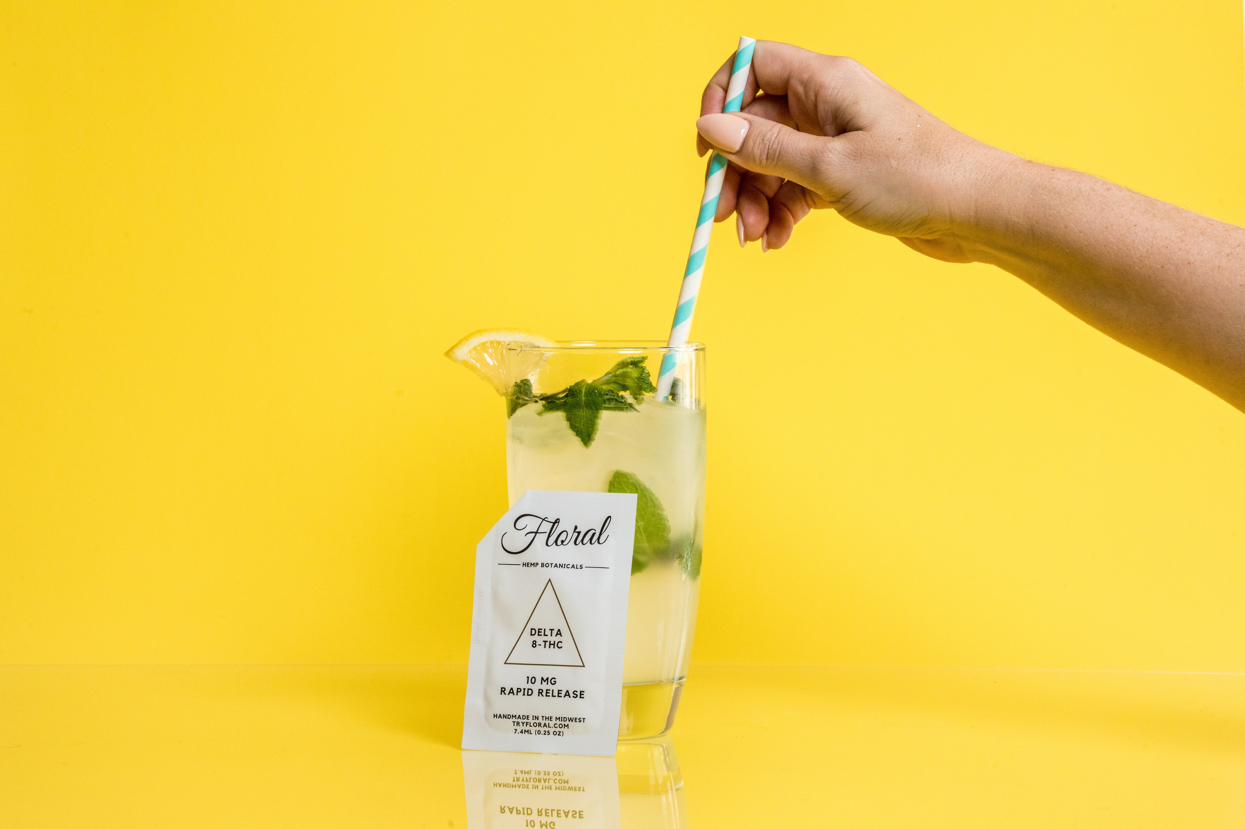 A hand stirring a packet of Floral Beverages' Floral Cocktail Enhancer into a cocktail glass against a bright yellow background.