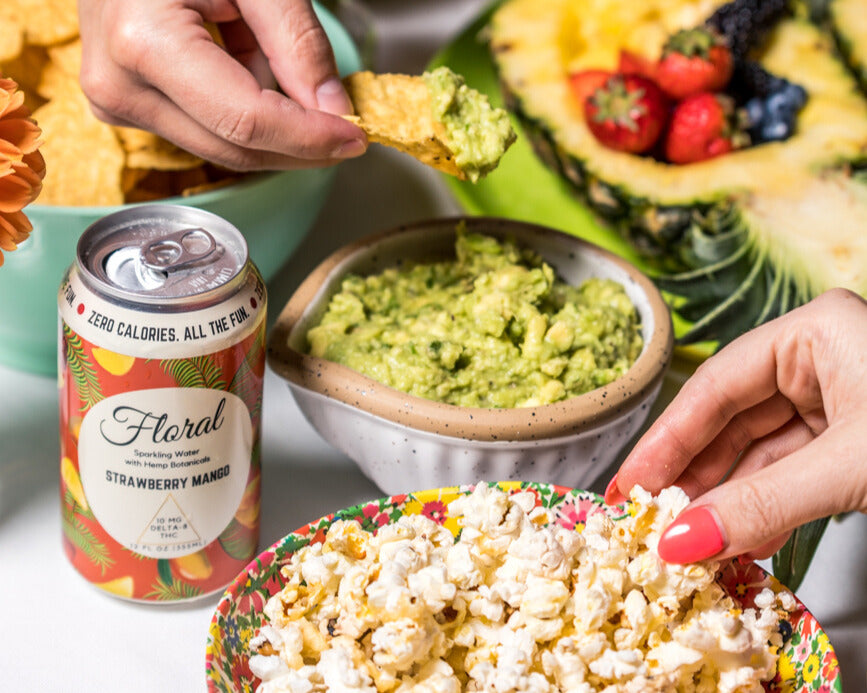 A can of Floral Beverages Strawberry Mango THC Seltzer sitting beside a bowl of guacamole and a bowl of popcorn at a party. 